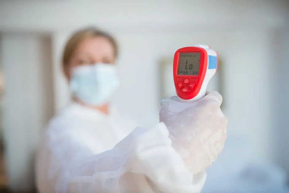 Masked healthcare worker using an infrared thermometer indoors for temperature screening.